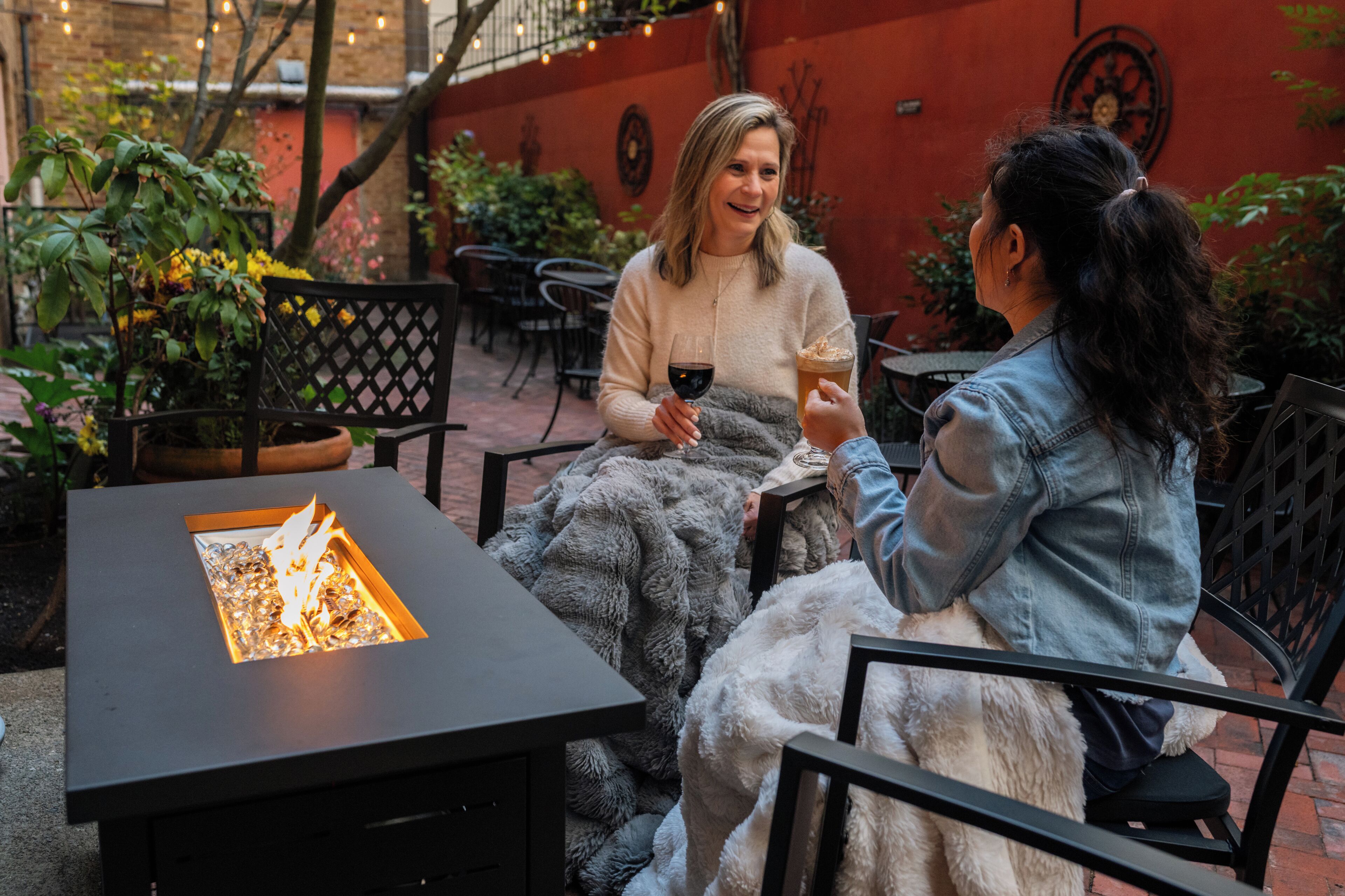 Dos personas sentadas en una mesa frente a una chimenea al aire libre, con plantas y un muro de ladrillo de fondo.