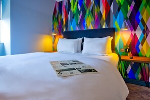 A neatly made bed with white linens and pillows, a colorful geometric-patterned wall behind, and a small table with a lamp beside the bed.