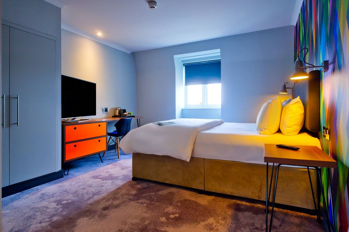 A bedroom with a bed, a desk, a chair, a TV, and a window.