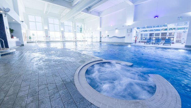 Une grande piscine intérieure avec un jacuzzi, un sol carrelé et un espace d’accueil doté de sièges.