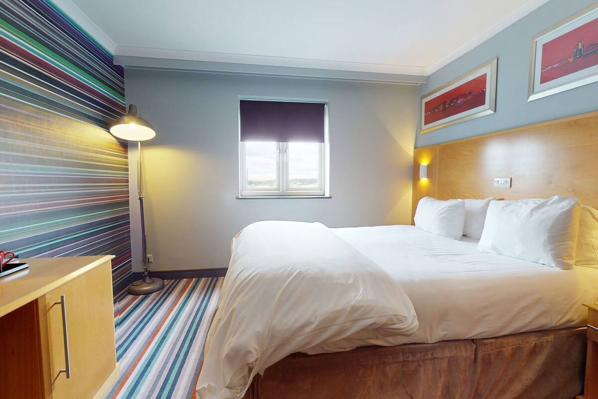 A hotel room with a bed, a desk, a lamp, a window, and a striped wall.
