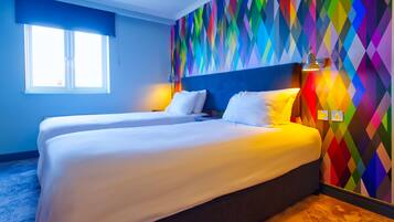 Habitación de hotel con dos camas, un muro con estampado de diamantes y colores, una ventana con persianas y una lámpara en la mesita de noche.