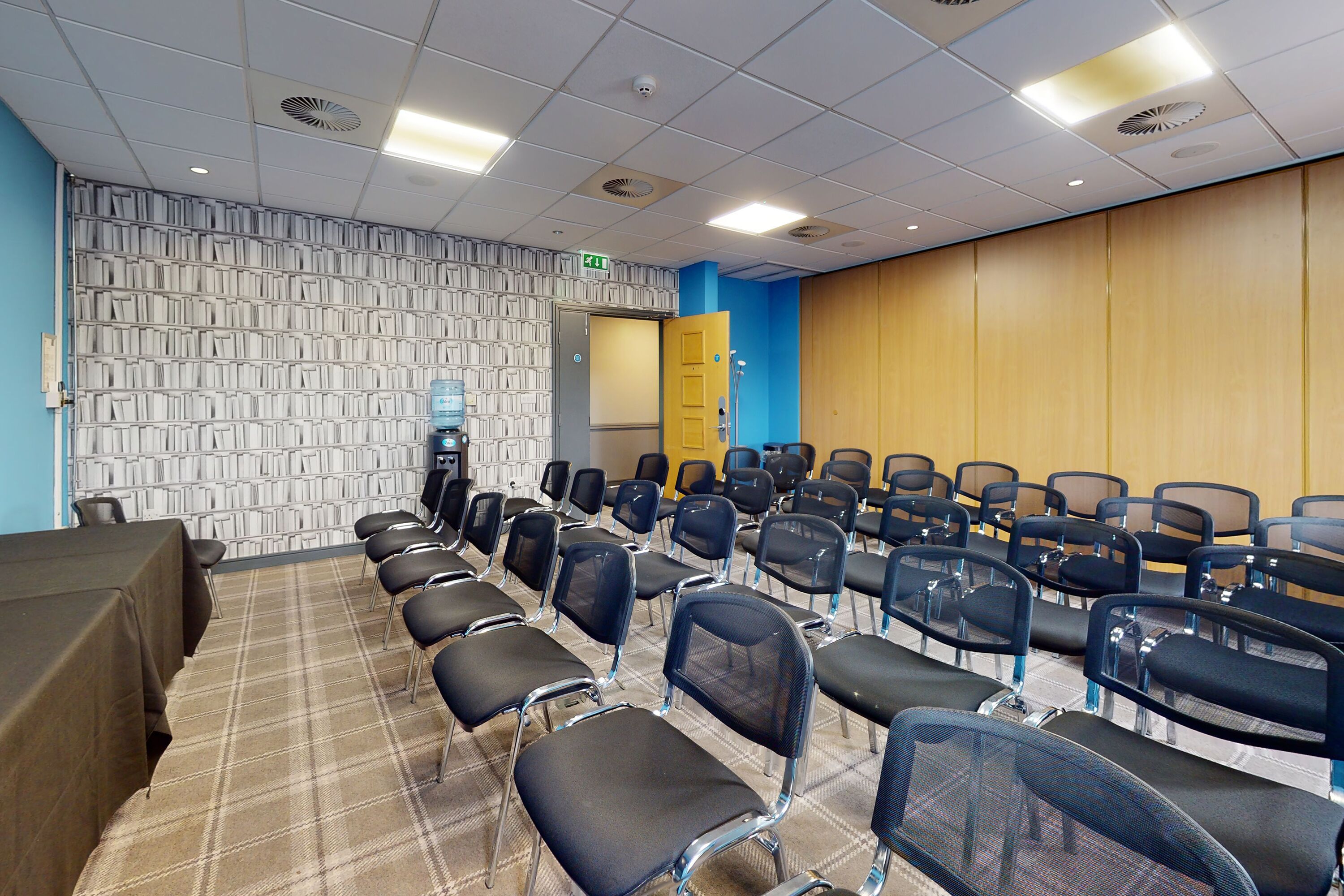 Un salón de conferencias con filas de sillas, una mesa, un dispensador de agua y una puerta.