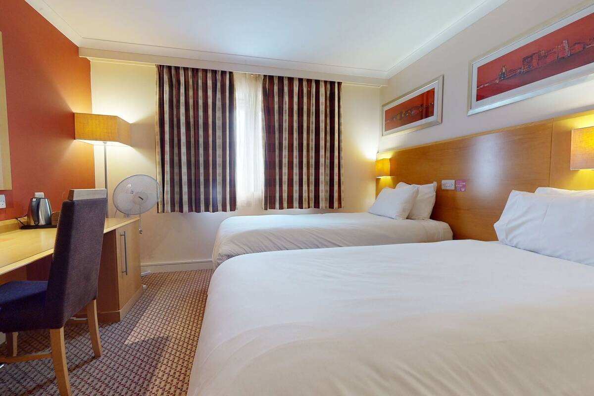 A hotel room with two beds, a desk, a chair, a fan, and a painting on the wall.