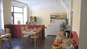 Un comedor con mesas y sillas de madera, una mesa de buffet con comida y bebidas, y una zona de cocina al fondo.