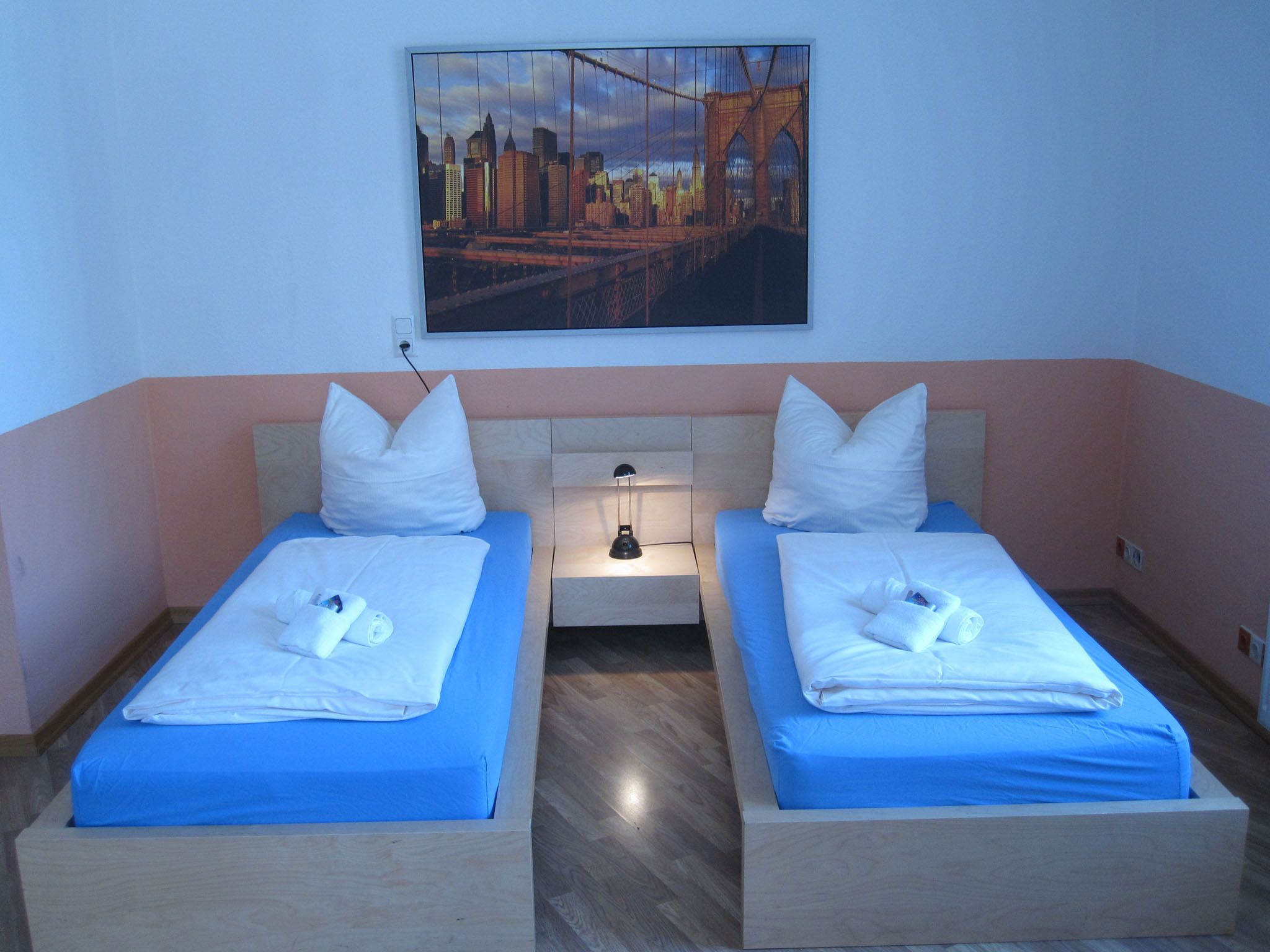 A hotel room with two beds, each with a blue mattress and white pillows, a nightstand with a lamp, and a cityscape painting on the wall.