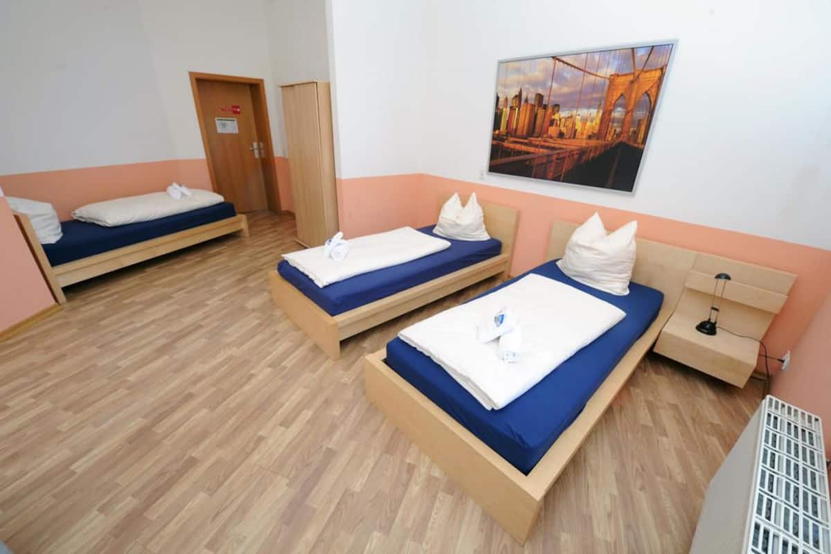 A room with three single beds, each with a blue mattress and white pillows. There is a wooden wardrobe and a small bedside table with a lamp. A cityscape painting is on the wall.