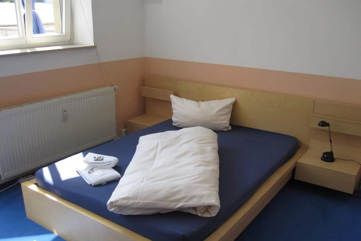 A single bed with a wooden frame, a blue mattress, and a white blanket. There is a white pillow, a folded white towel, and a bedside table with a lamp.