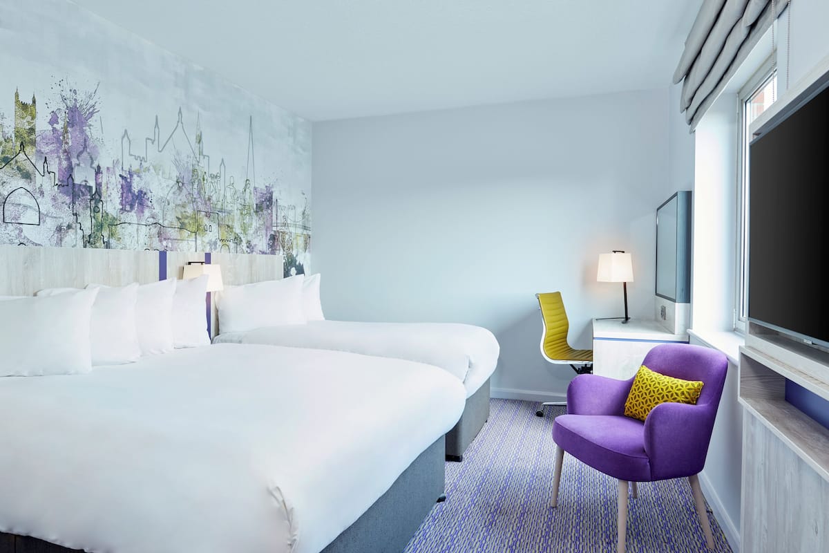 A hotel room with a large bed, a purple armchair, a television, and a cityscape mural on the wall.