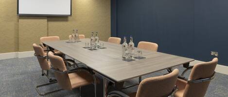 Un salón de conferencias con una larga mesa, sillas color beige y una pantalla para proyector.