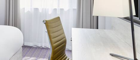 A modern hotel room with a curved upholstered chair, a white bed, a wooden desk with a lamp, and a view of the city through sheer curtains.