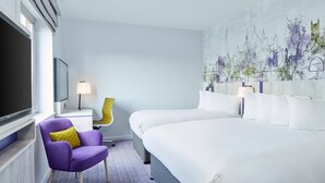 Habitación de hotel con una cama grande, un sillón púrpura con un cojín amarillo, un escritorio con una lámpara y un mural de paisaje urbano en la pared.