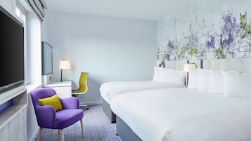 A hotel room with a large bed, a purple armchair with a yellow pillow, a desk with a lamp, and a cityscape mural on the wall.