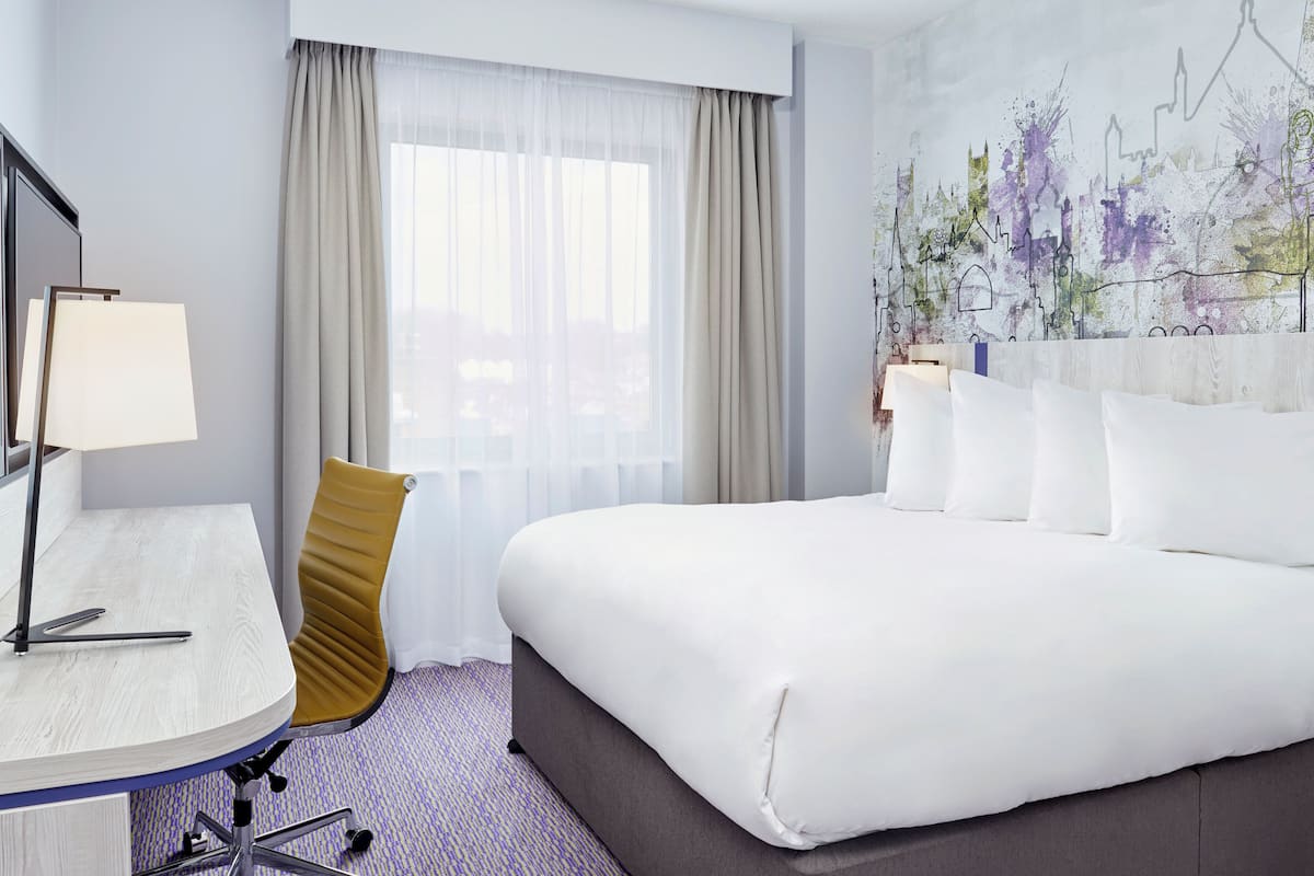 A hotel room with a large bed, a desk with a lamp, a chair, and a window with sheer curtains.