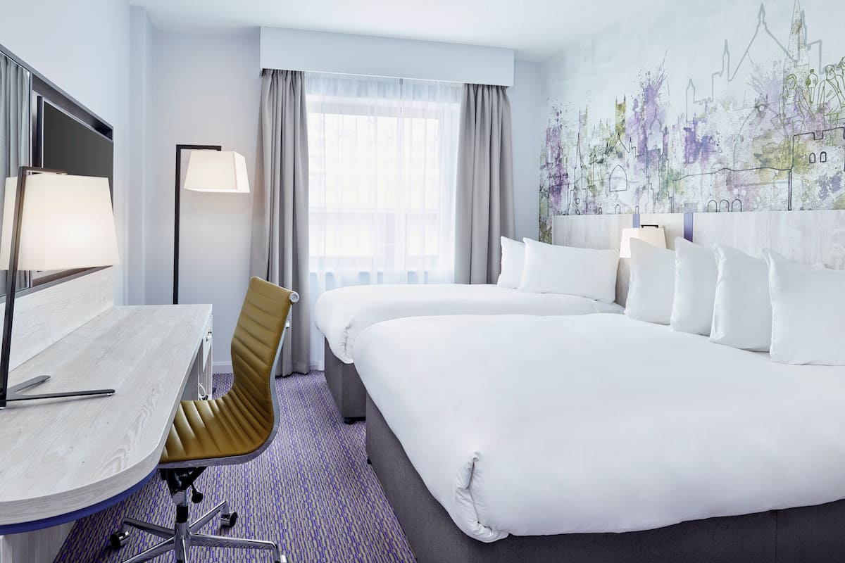 A hotel room with a bed, a desk with a lamp, a chair, and a large window with curtains.
