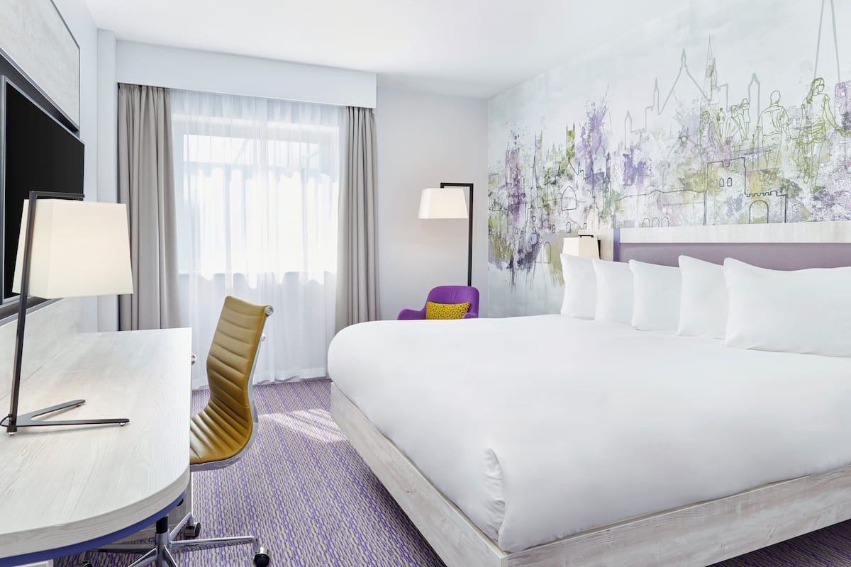 A hotel room with a large bed, a desk with a lamp, a chair, and a painting on the wall.