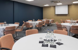 A conference room with round tables, chairs, water bottles, glasses, and a projector screen.