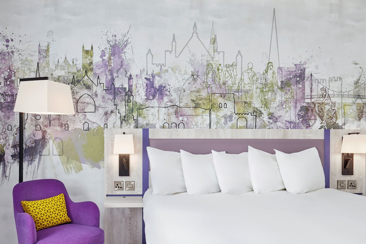 A hotel room with a large bed, a purple armchair, and a cityscape mural on the wall.