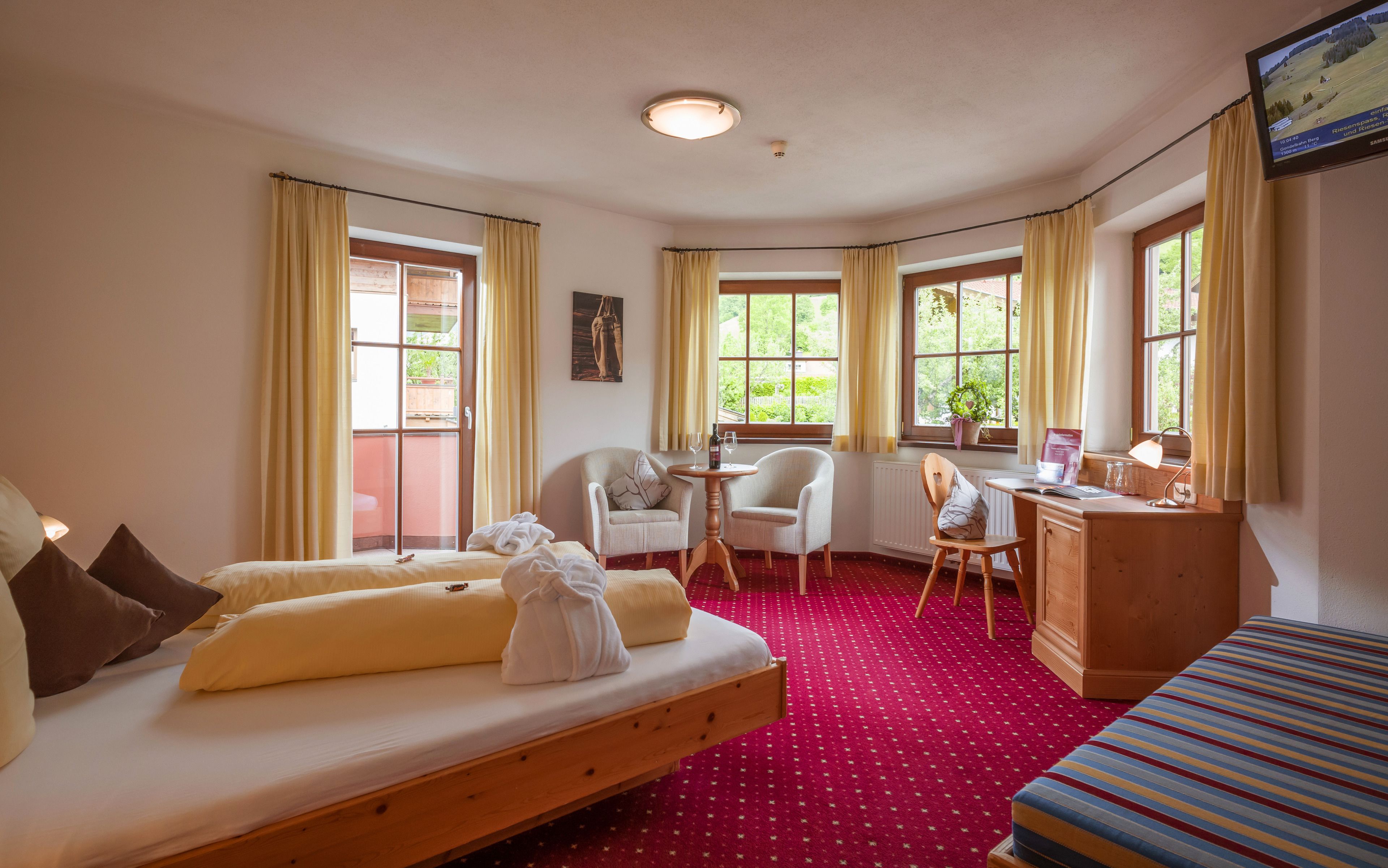 Una habitación de hotel con cama, escritorio, silla y sofá. Hay ventanas con cortinas, un radiador y un televisor en la pared.