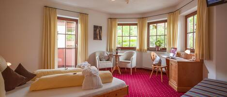 Una habitación de hotel con cama, escritorio, silla y sofá. Hay ventanas con cortinas, un radiador y un televisor en la pared.