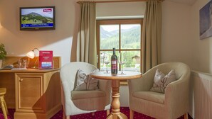 A room with two armchairs, a wooden table with a bottle and glasses, a TV on the wall, and a window with a view of mountains. - Hotel Reitlwirt (Brixen im Thale)