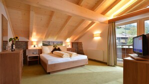 A bedroom with a wooden ceiling, a bed, a nightstand, a dresser, a TV, and a window with a view of a house and trees. - Hotel Reitlwirt (Brixen im Thale)