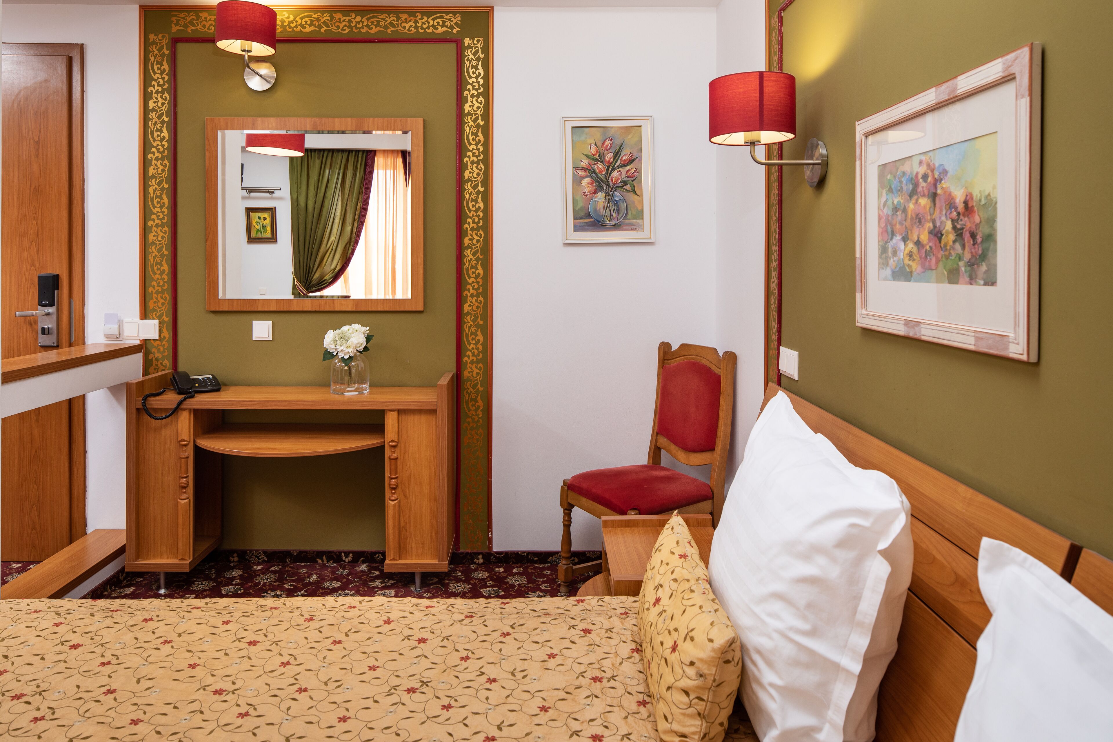 Habitación de hotel con escritorio de madera, una silla, cama con almohadas blancas y estampadas, espejo con cortina verde y dos lámparas de pared.