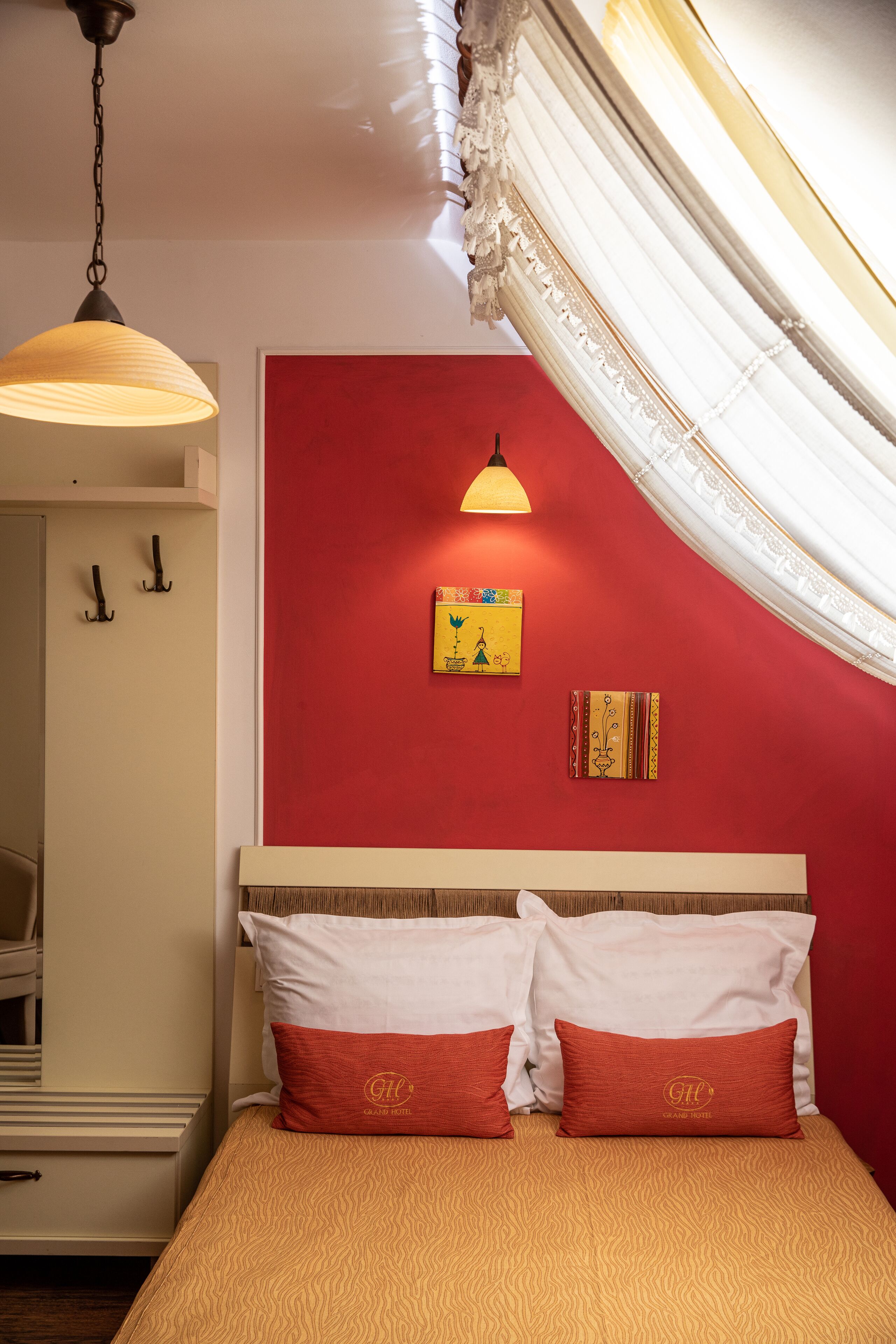A hotel room with a red accent wall, a bed with white and red pillows, a hanging lamp, and a decorative wall hanging.