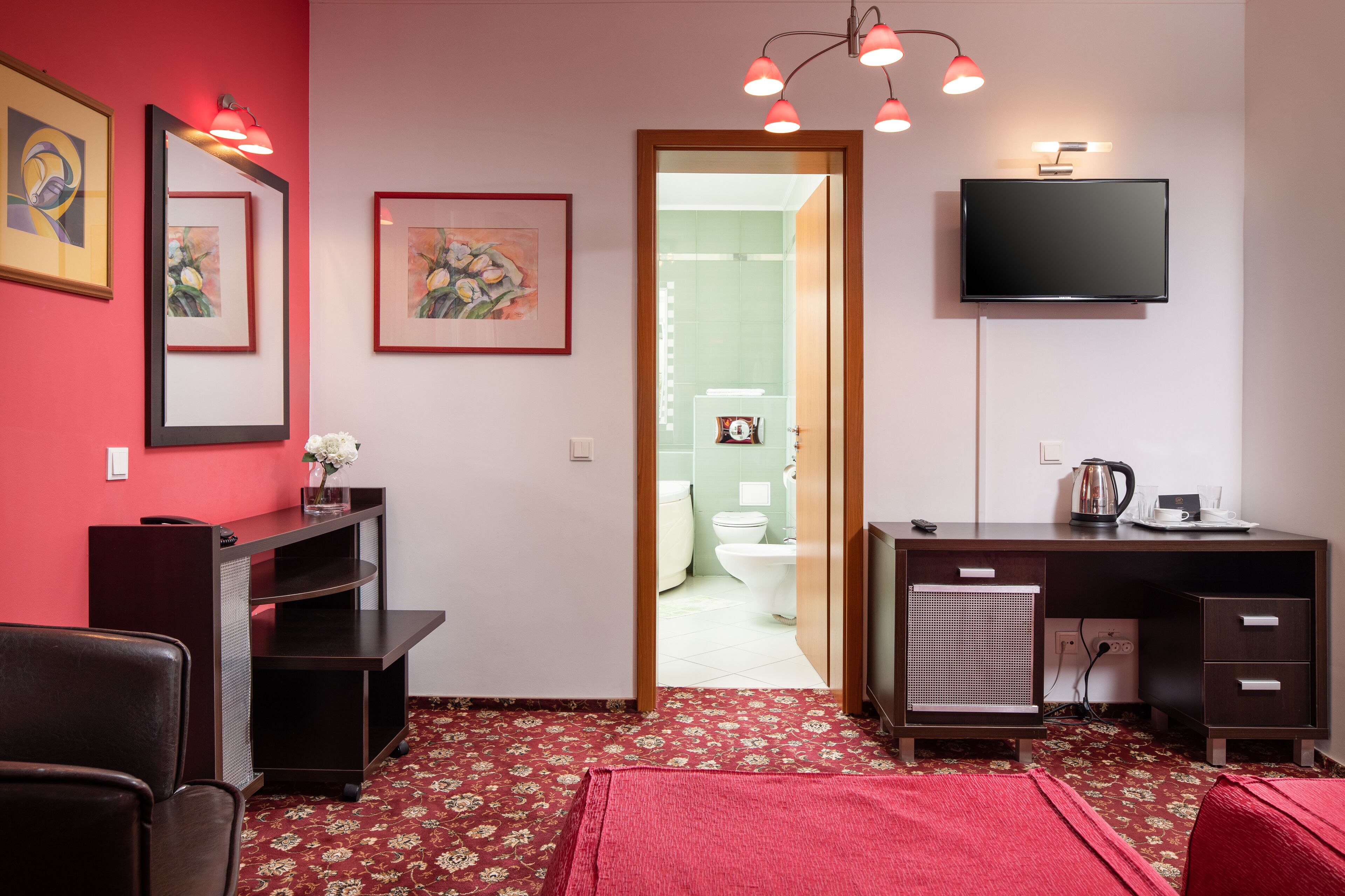 A compact hotel room with a red carpet, a desk with a flat-screen TV, a small kitchenette, and a door leading to a bathroom.