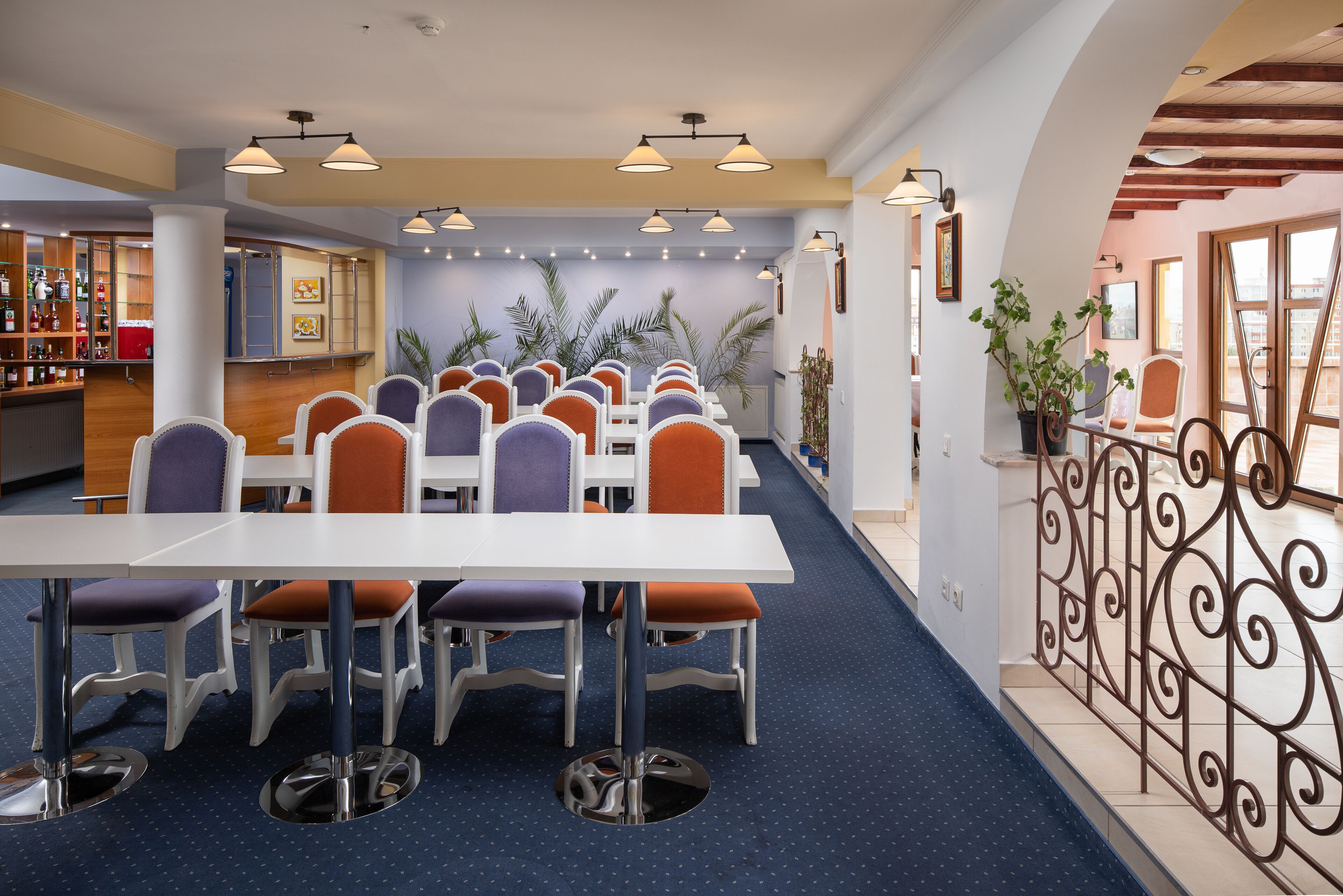 Un comedor con varias mesas y sillas, una zona de bar con estantes llenos de botellas, y una escalera con barandilla de hierro forjado.