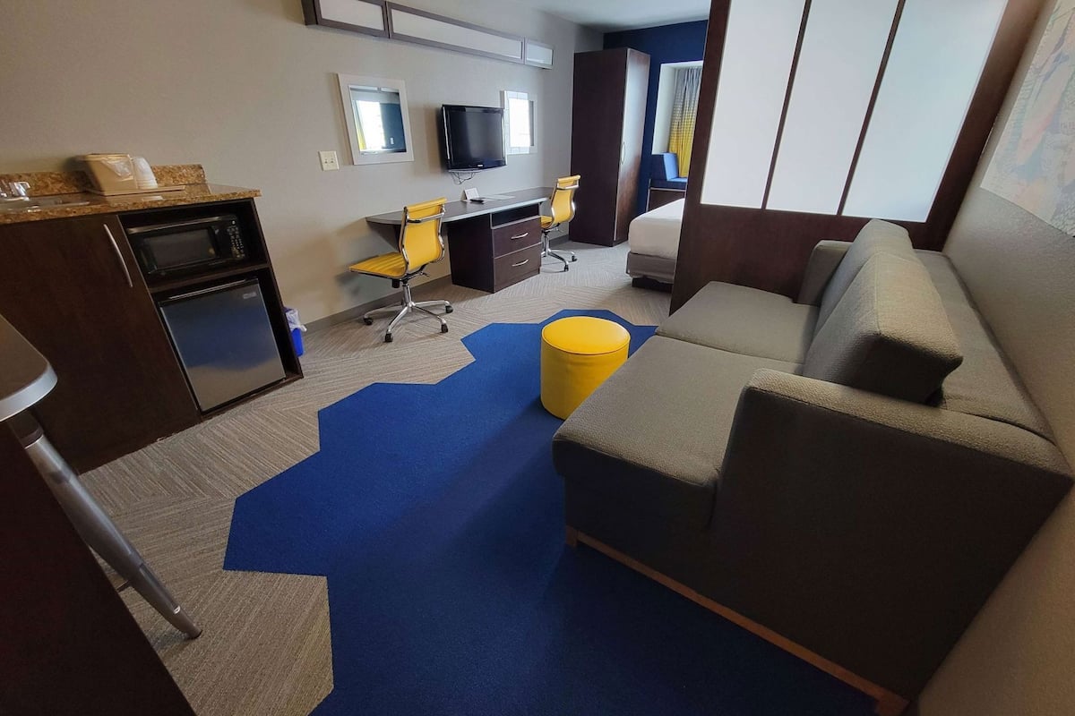 A hotel room with a kitchenette, a sofa, a desk with a chair, a TV, and a bed.