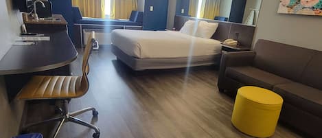 Habitación de hotel con cama, escritorio con silla, sofá, reposapiés amarillo y un cubo de basura.