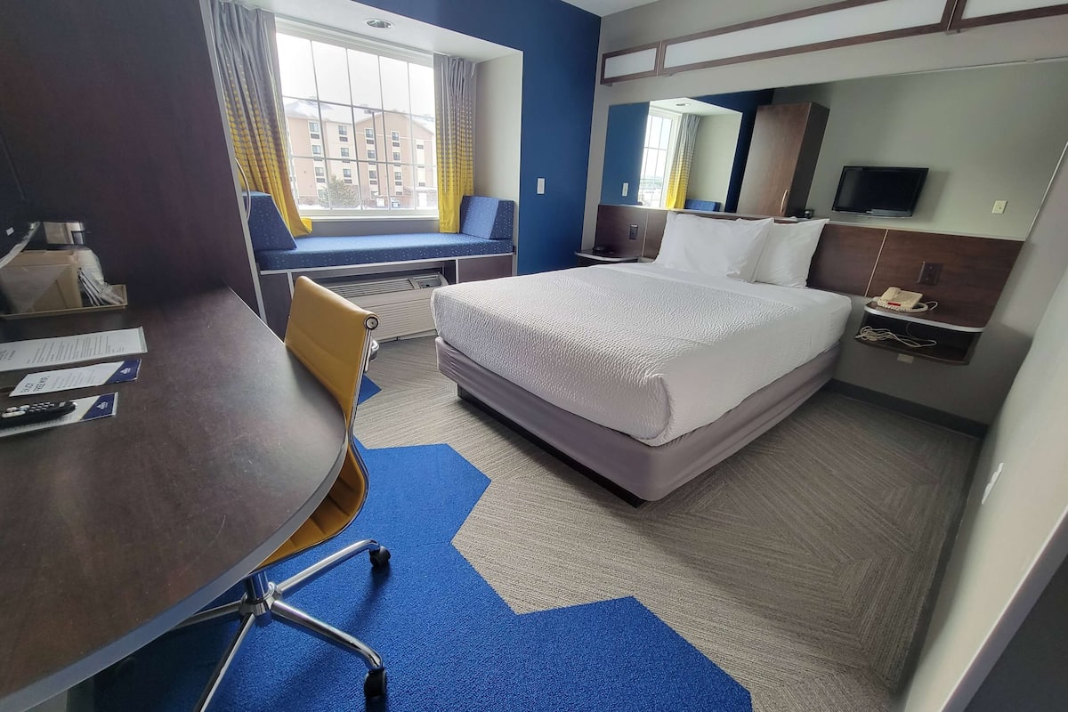 A hotel room with a bed, a desk, a chair, a window with a view of buildings, a television, and a telephone.