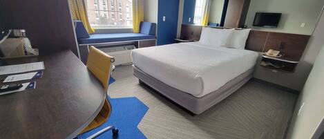 A hotel room with a bed, a desk, a chair, a window with a view of buildings, a television, and a telephone.