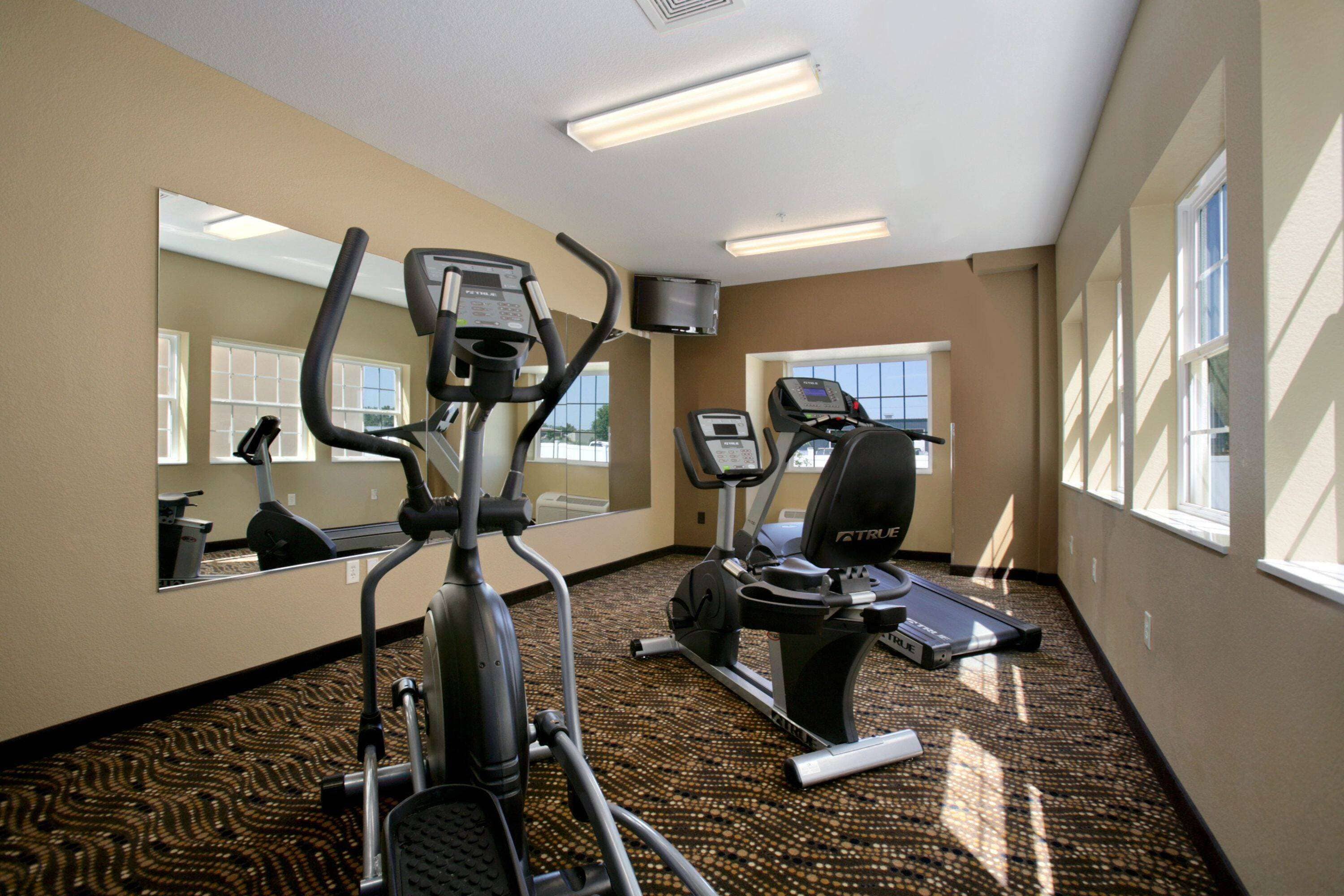 A hotel fitness room with exercise equipment, a large mirror, and a carpeted floor.