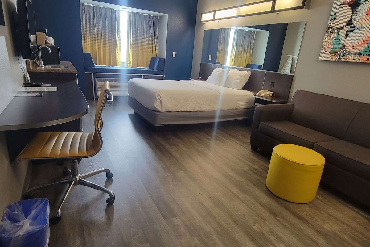 A hotel room with a bed, a desk, a chair, a sofa, a yellow ottoman, a mirror, and a window with curtains.