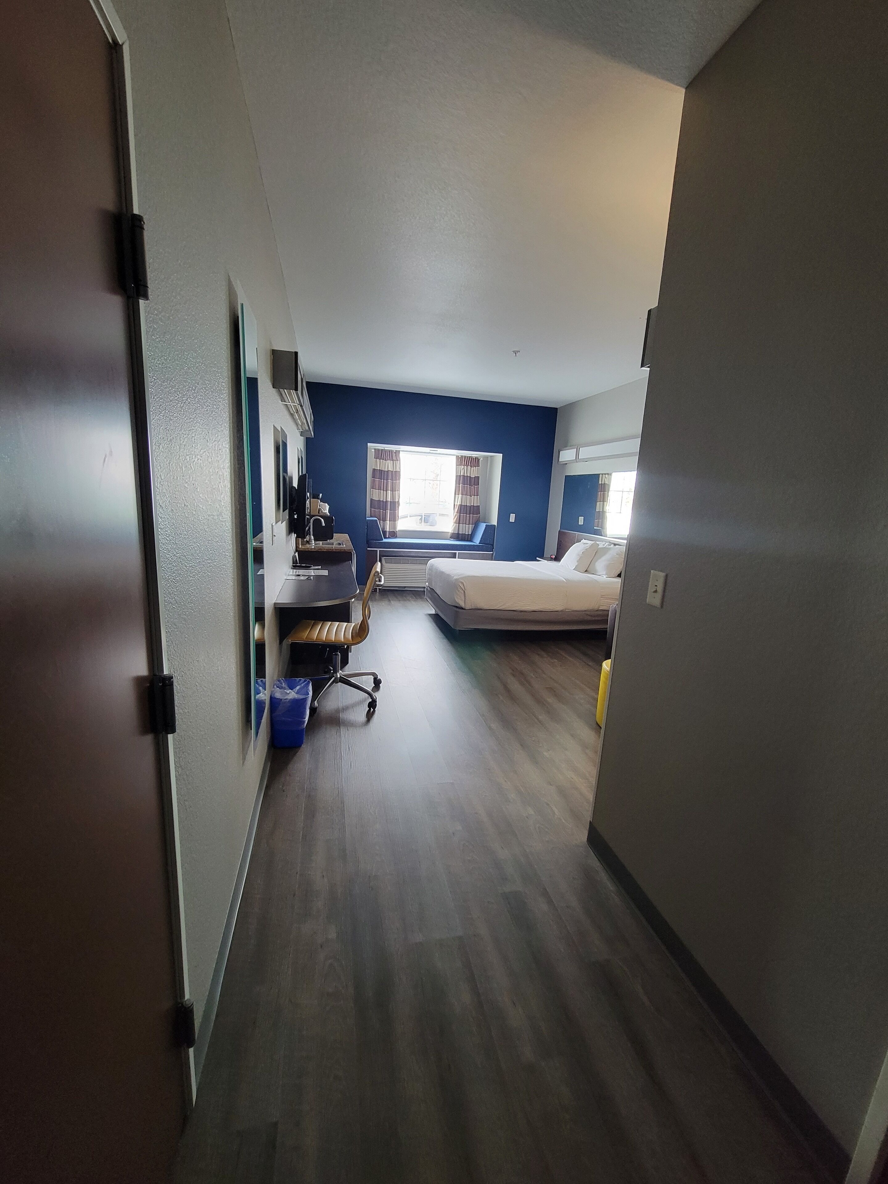 A hotel room with a bed, a desk with a chair, a window with curtains, and a blue wall.