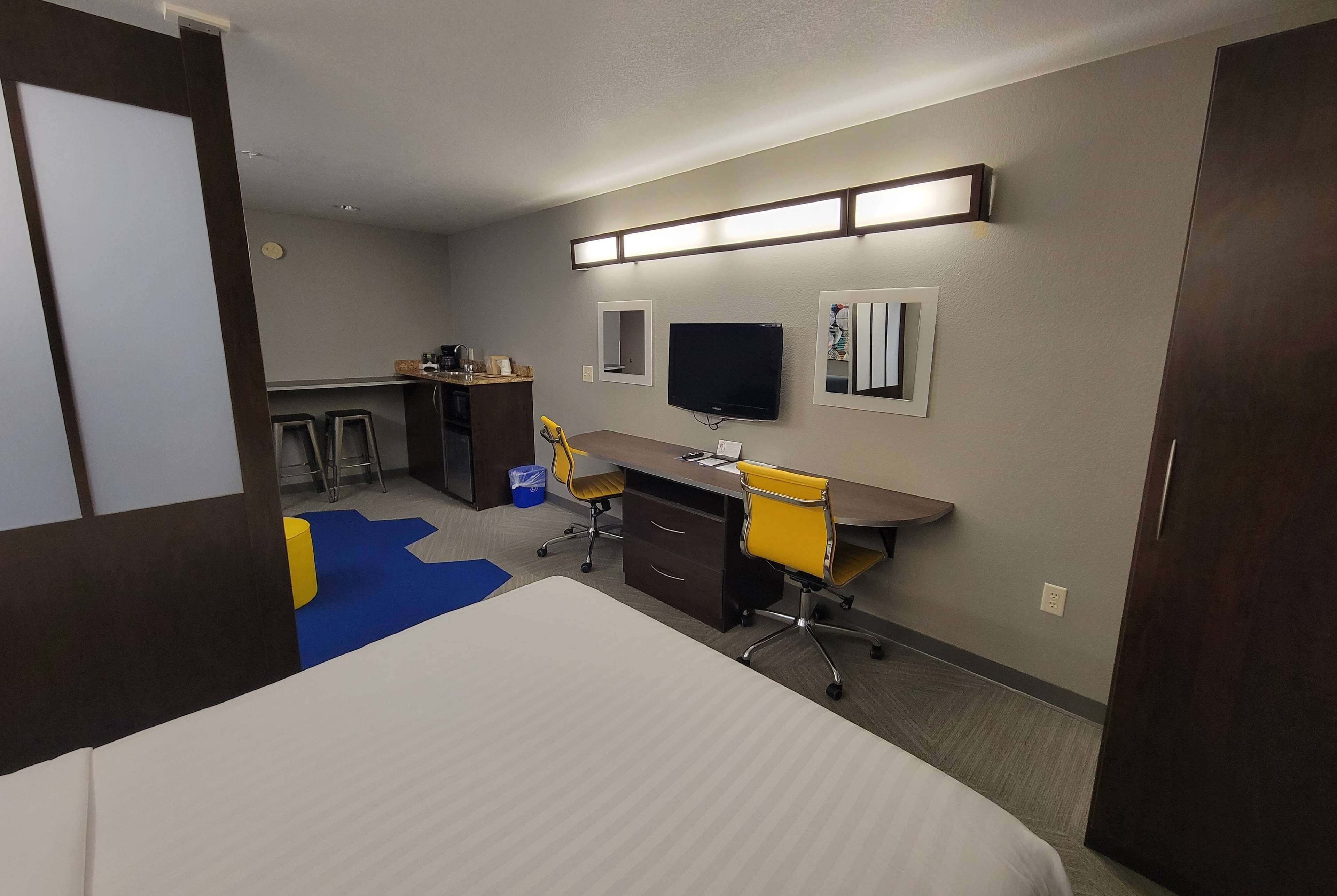 A hotel room with a bed, desk, chair, TV, and a small kitchenette area.