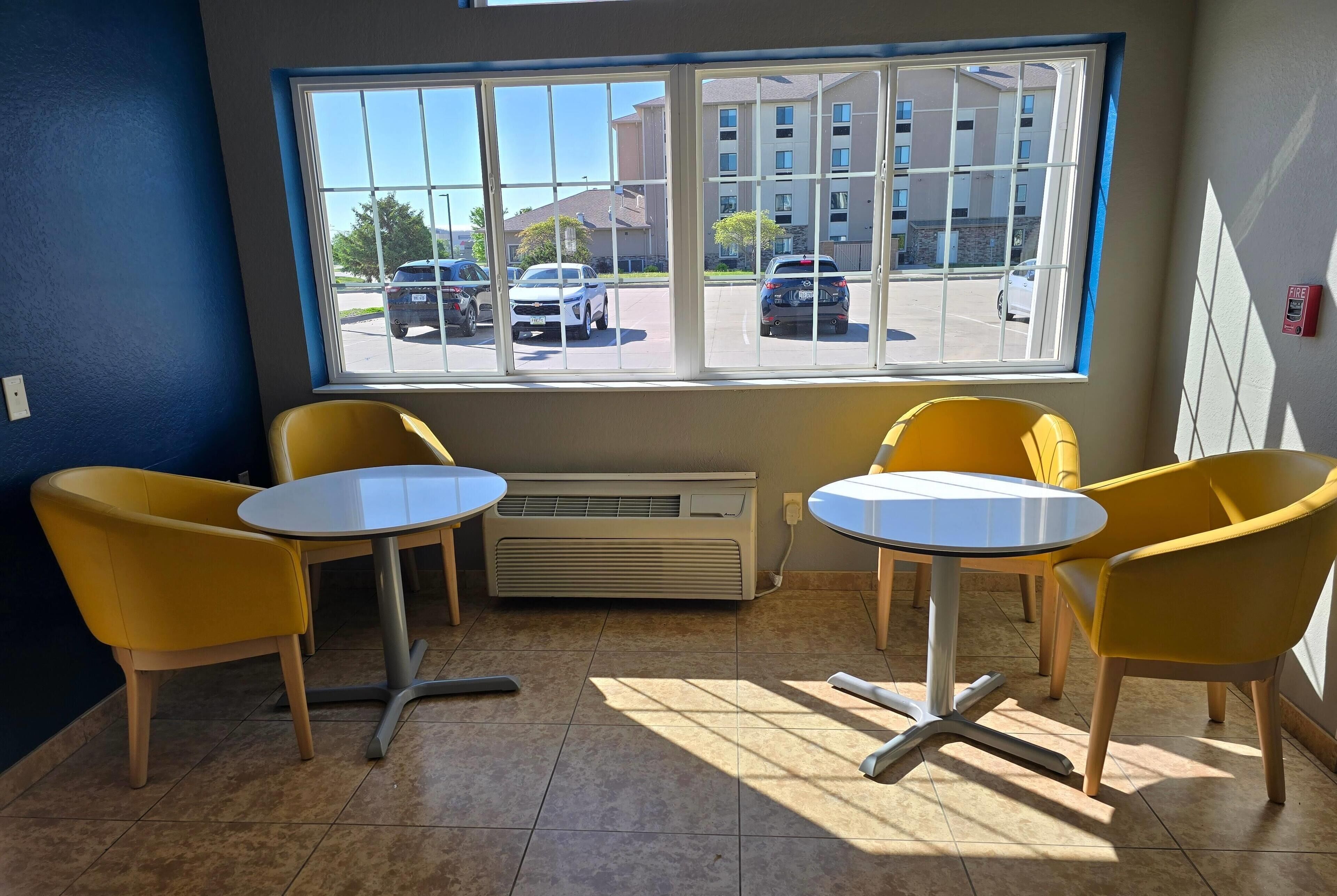 Une pièce avec deux chaises jaunes et une petite table, une fenêtre donnant sur un parking et des immeubles, et un climatiseur fixé au mur.