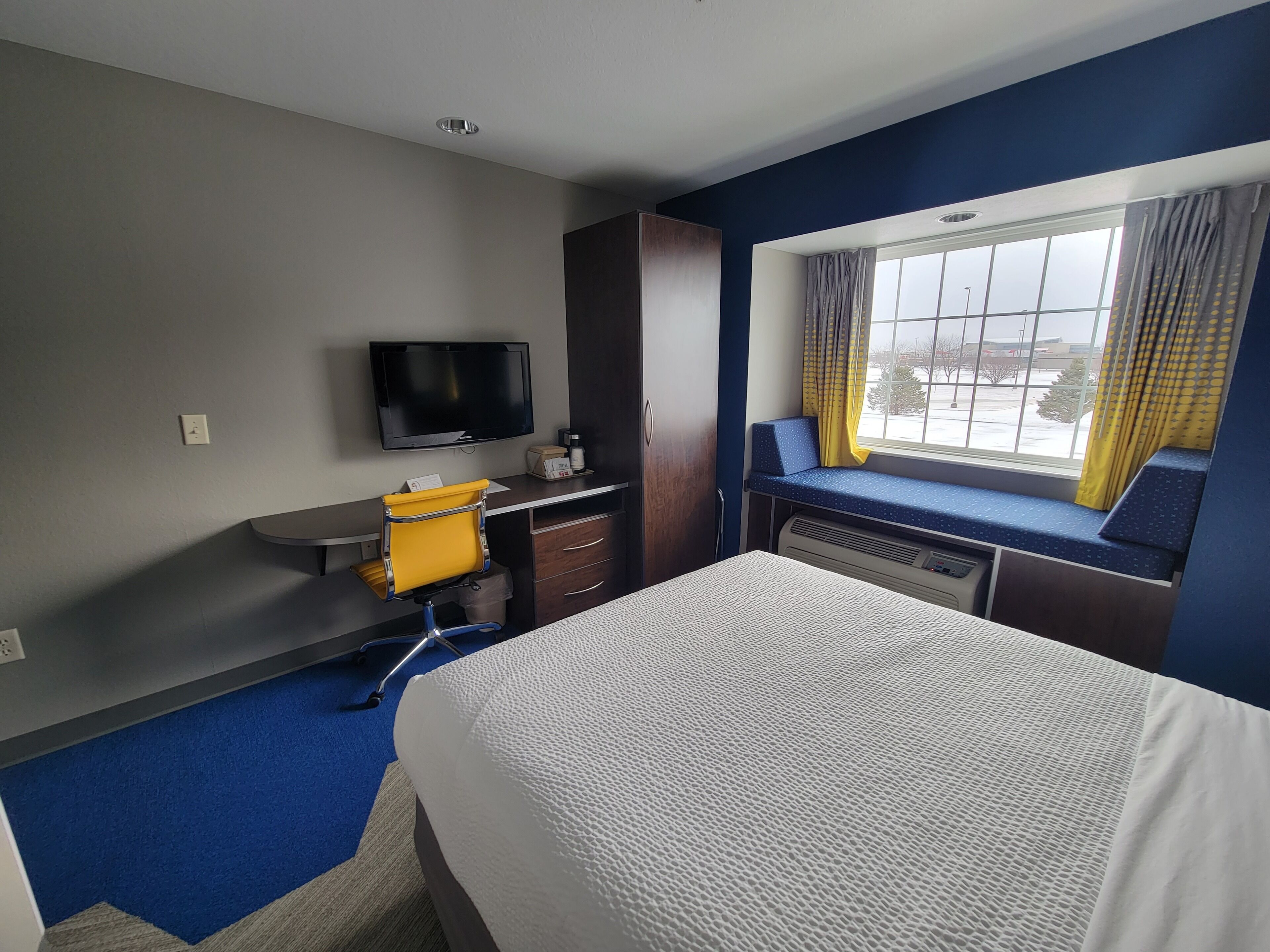 Une chambre d’hôtel avec un lit, un bureau et une chaise, une télévision et une fenêtre avec des rideaux.