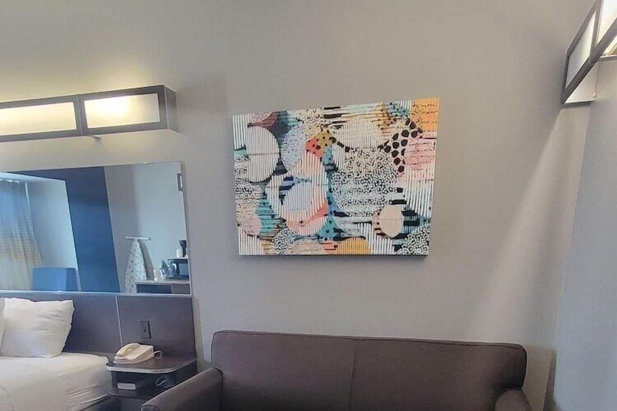 A modern hotel room with a brown sofa, a yellow ottoman, a bed with white bedding, a nightstand with a lamp, and a colorful abstract painting on the wall.