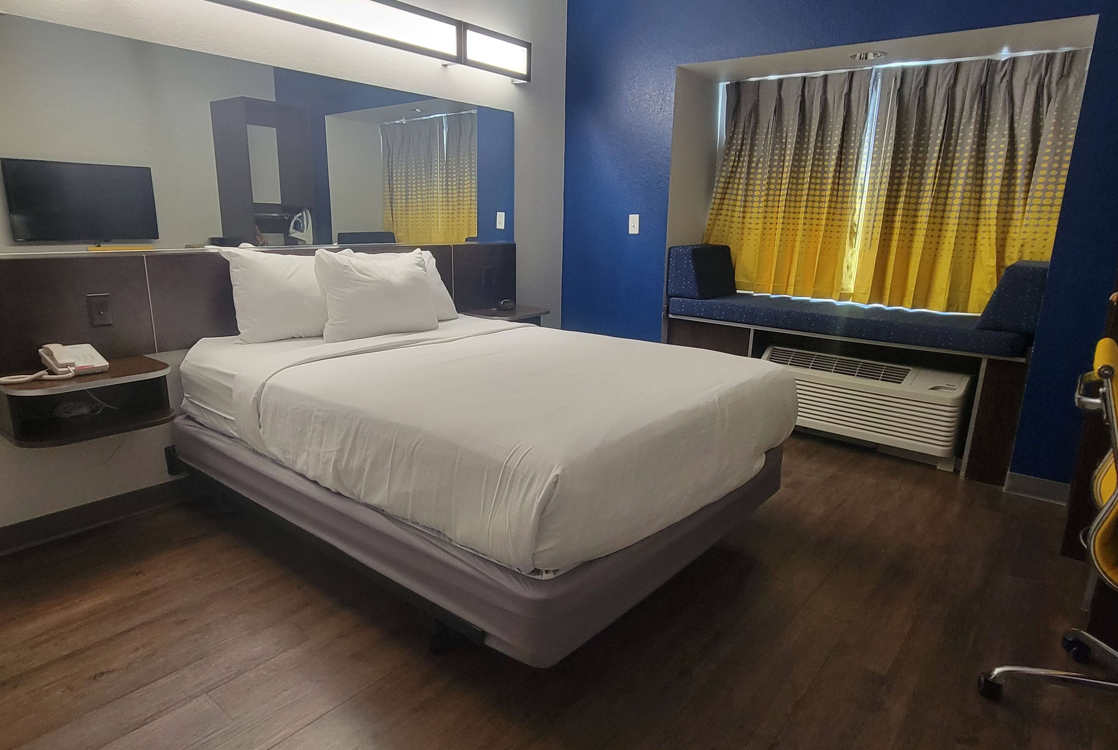 A hotel room with a large bed, a bedside table with a phone, a mirror, a TV, and a window with curtains.