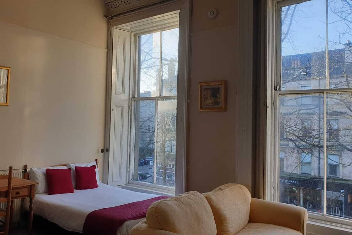 A room with a bed, a wooden chair, a table, and a window with a view of buildings.
