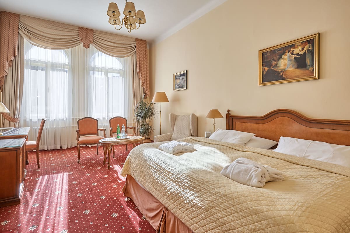 Ein Hotelzimmer mit einem großen Bett, einem Schreibtisch, einem Stuhl und einem kleinen Tisch. An der Wand hängt ein Bild und es gibt ein Fenster mit Vorhängen.