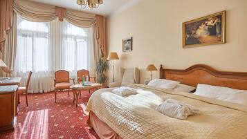 Una camera d'albergo con un letto grande, una scrivania, una sedia e un tavolino. Si vede un quadro appeso al muro e una finestra con tende.