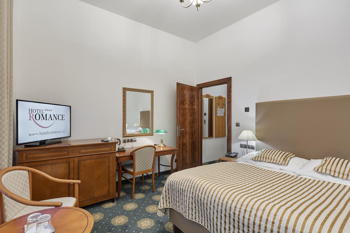 Ein Hotelzimmer mit einem Bett, einem Schreibtisch, einem Stuhl, einem Fernseher, der 'HOTEL ROMANCE' zeigt, und einem Spiegel.