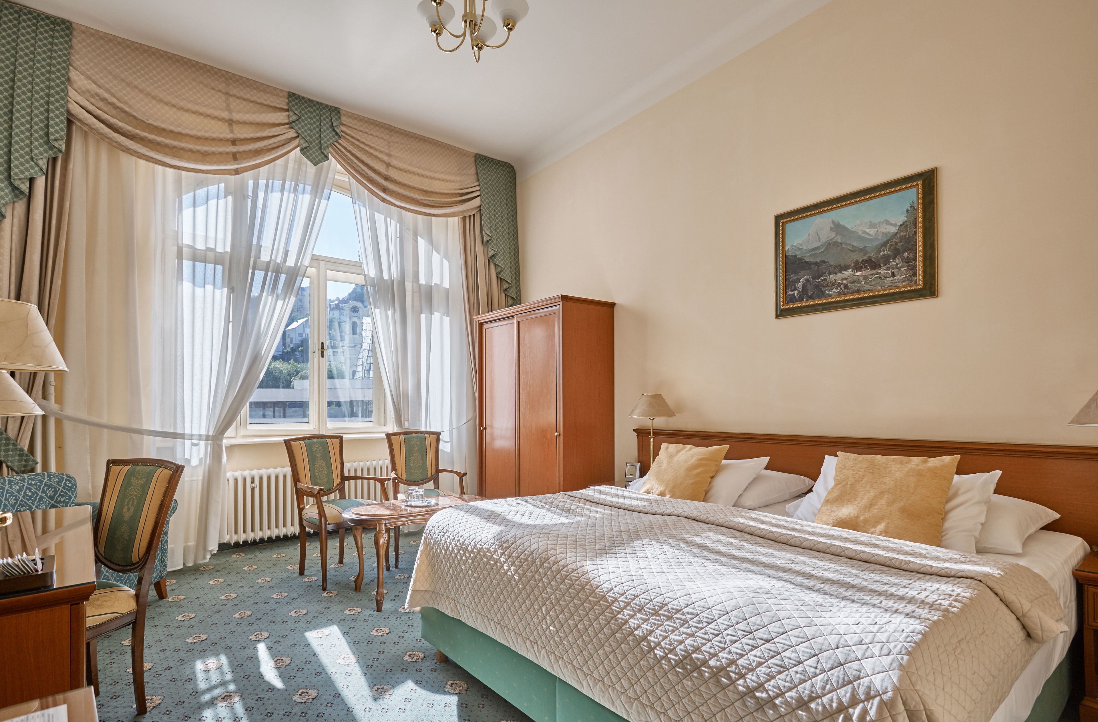 A hotel room with a bed, a chair, a table, a radiator, and a painting on the wall.