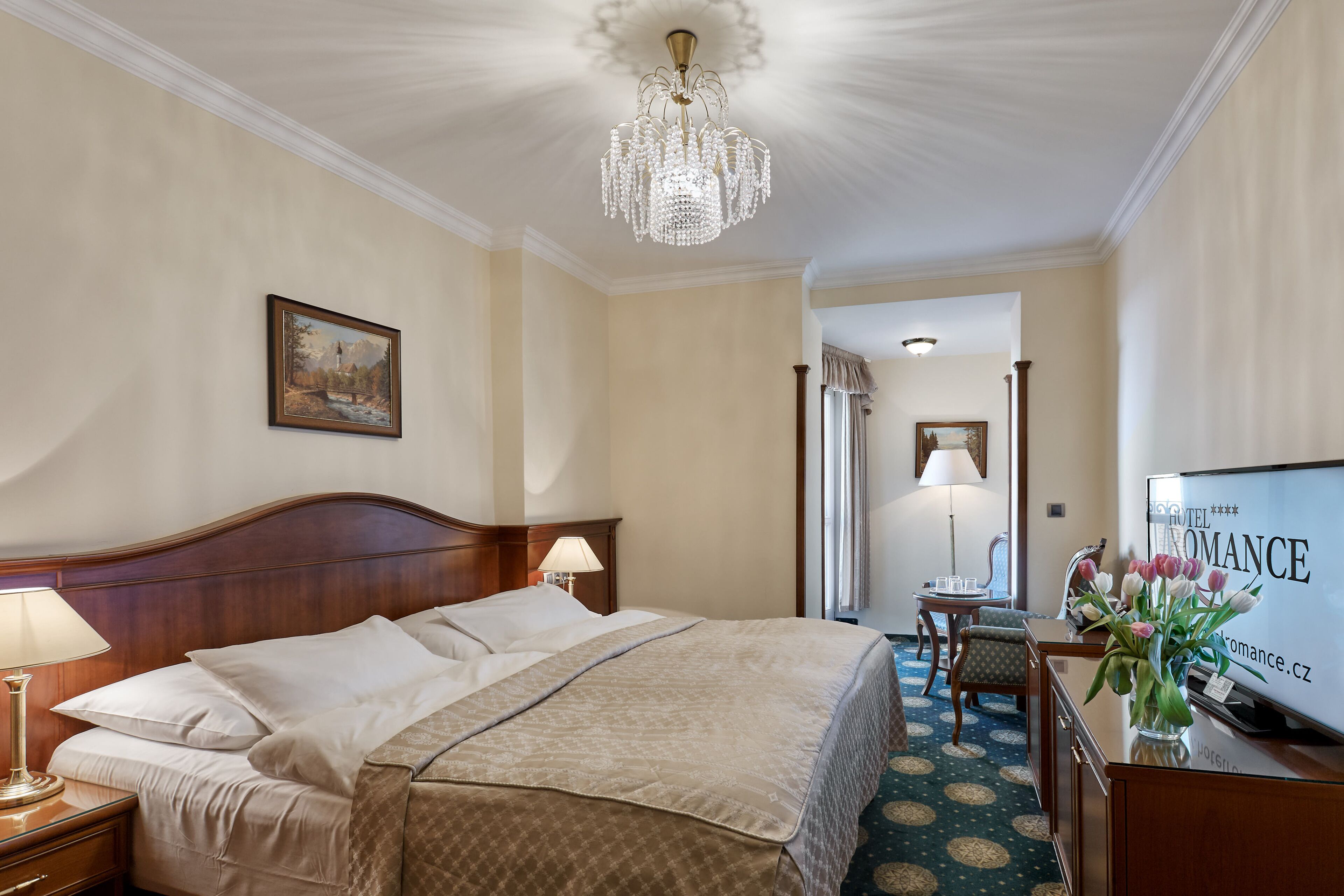 Ein Hotelzimmer mit einem großen Bett, einer Chandelüster, einem Schreibtisch mit Computer und einem Fernseher.