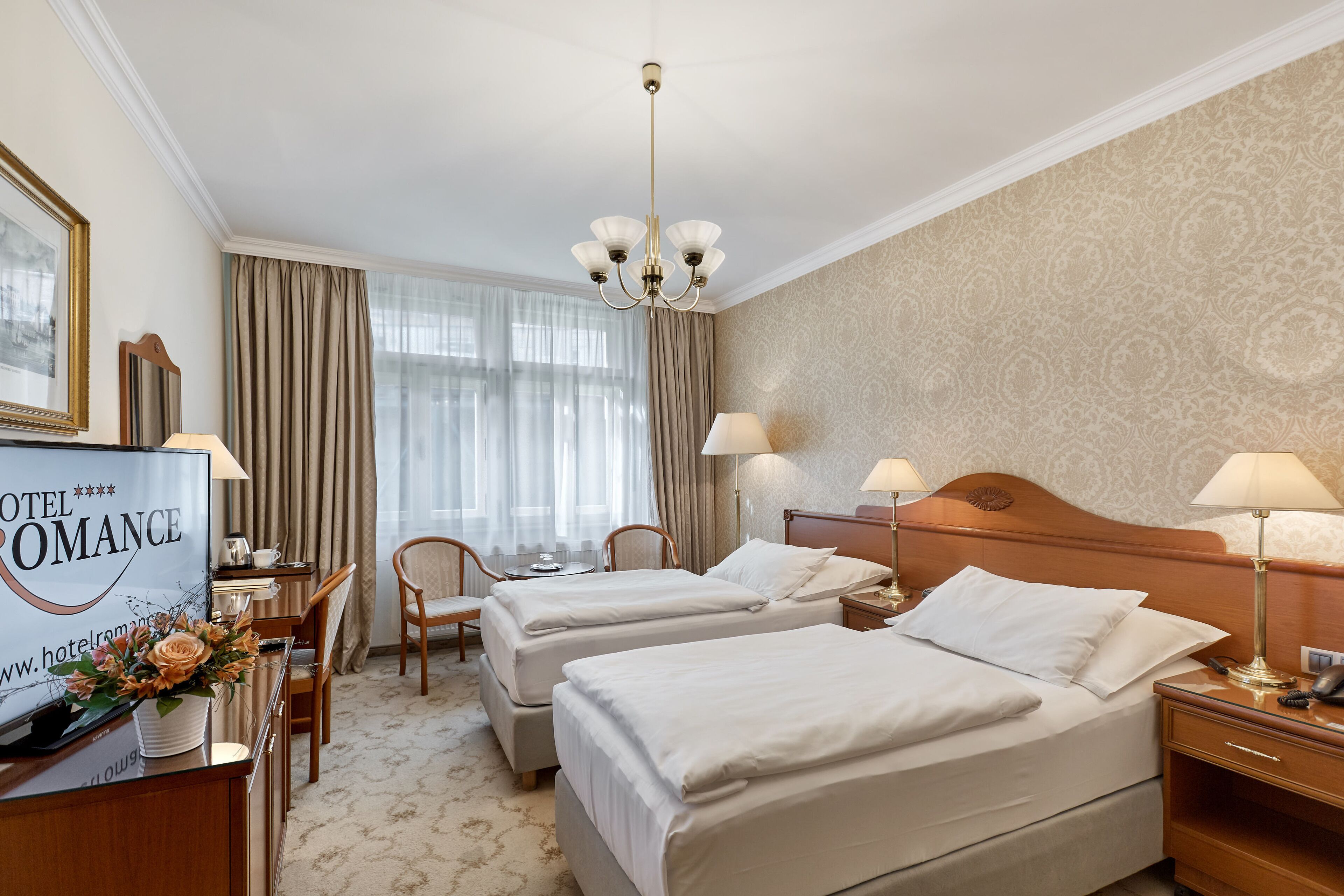Quarto de hotel com duas camas, uma escrivaninha com televisão, uma cadeira e uma luminária de mesa.