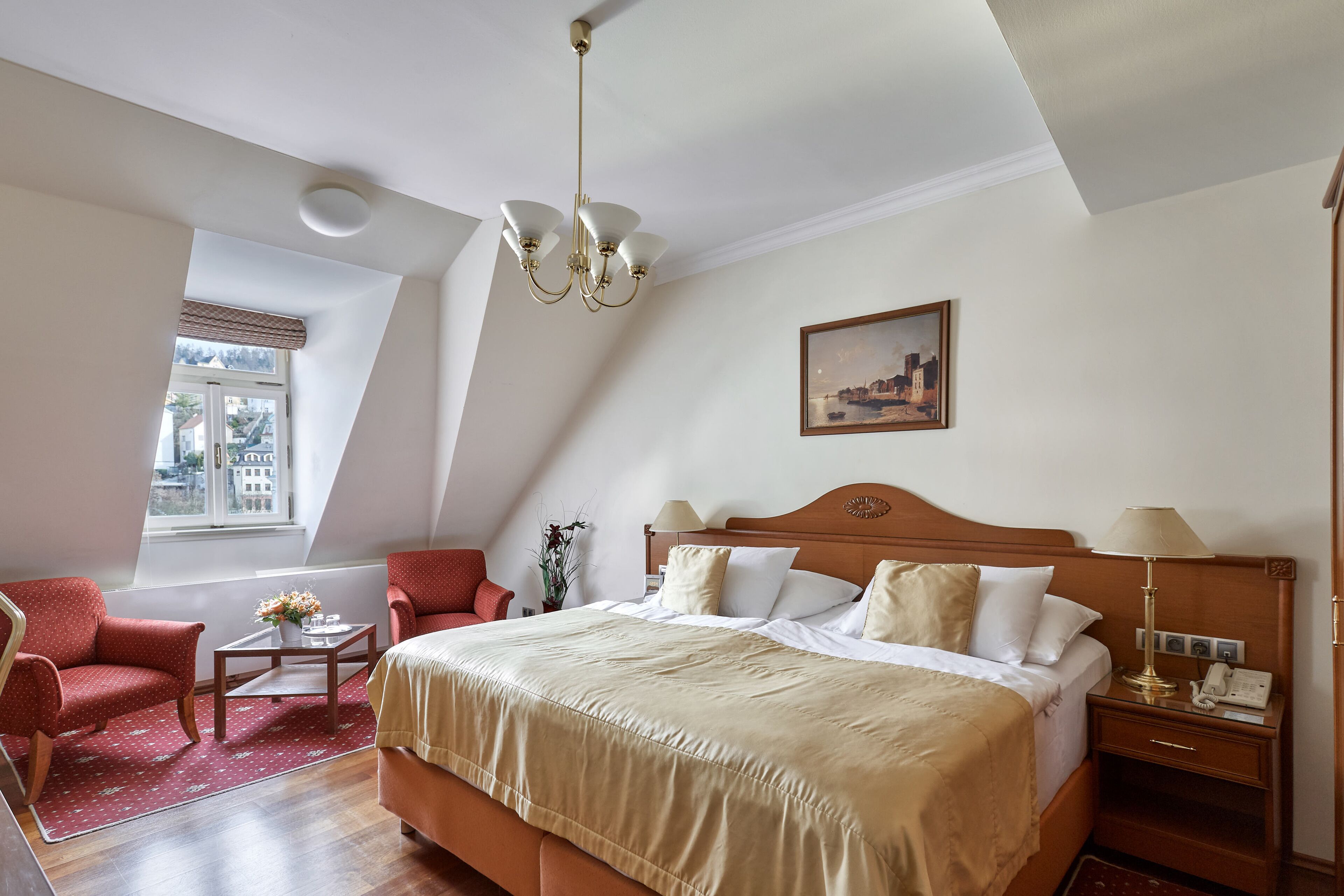 Una habitación de hotel con una cama grande, una mesita de noche de madera, un sillón rojo, una pequeña mesa con un jarrón de flores y un cuadro en la pared.